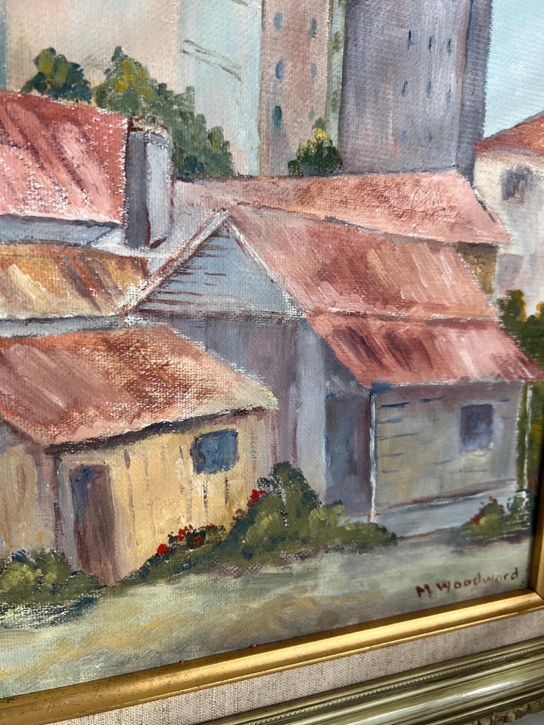 May include: Oil painting of a European village scene with red-tiled roofs and light-colored buildings. The artwork is framed in a gold-colored frame and signed by M. Woodward. The painting depicts a sunny day with a clear sky.