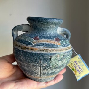 May include: A small, blue ceramic vase with two handles. The vase has a textured surface with painted details, including a fish, bamboo, and a landscape scene. A tag is attached.