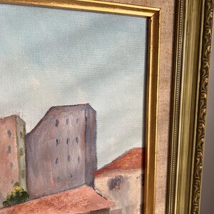 May include: An oil painting of a cityscape with buildings and rooftops in shades of red, brown, and gray. The painting is framed in a gold-colored ornate frame.