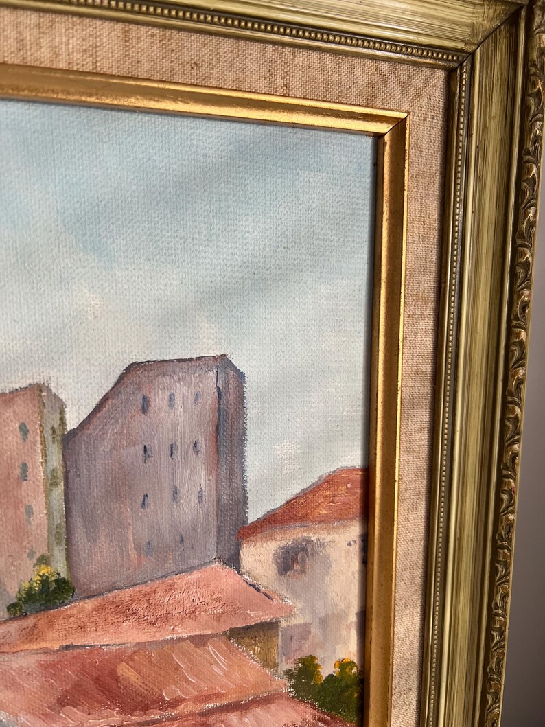 May include: An oil painting of a cityscape with buildings and rooftops in shades of red, brown, and gray. The painting is framed in a gold-colored ornate frame.