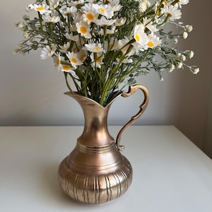 May include: A metallic brass-toned pitcher with a curved handle and a flared rim, filled with a bouquet of white daisy-like flowers featuring yellow centres and green stems. The pitcher has a rounded body with vertical ribbed detailing.