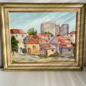May include: Oil painting of a cityscape with buildings and red-roofed houses, set against a light blue sky. The artwork is framed in a gold-colored, ornate frame. Signed by M. Woodward.