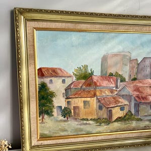 May include: Oil painting of a cityscape with buildings and trees, framed in a gold ornate frame. The painting features buildings with red roofs and a light blue sky. The frame has a gold finish.