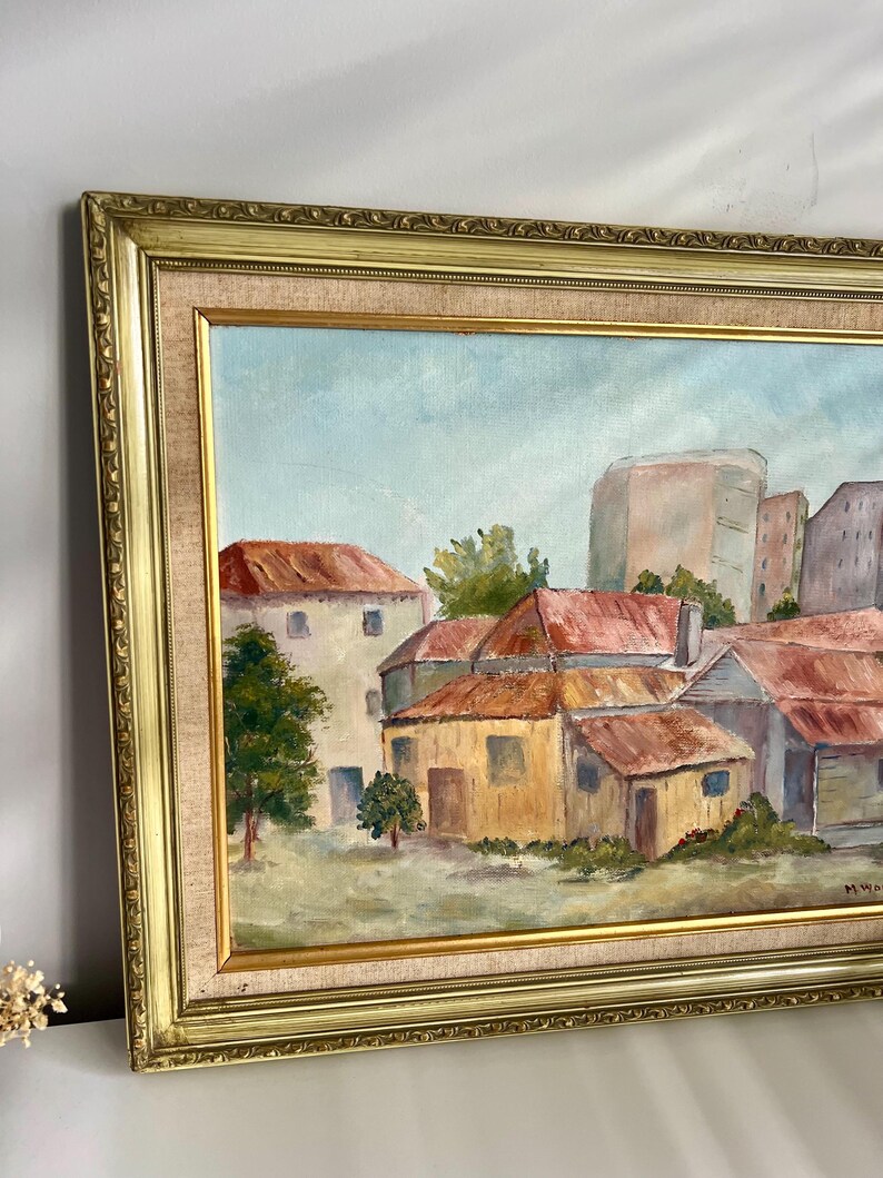 May include: Oil painting of a cityscape with buildings and trees, framed in a gold ornate frame. The painting features buildings with red roofs and a light blue sky. The frame has a gold finish.
