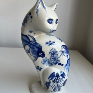 May include: A white ceramic cat figurine decorated with blue underglaze patterns. The design features detailed illustrations of swimming fish, pine needles, and lotus flowers across the body, with blue painted eyes and facial markings.