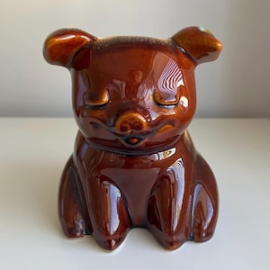 May include: A glossy, dark brown ceramic figurine shaped like a seated pig. The pig has a smooth, reflective surface, rounded ears, and a small snout. Its eyes are depicted as closed with simple, curved lines, giving the animal a peaceful expression.