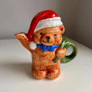 May include: A ceramic teddy bear figurine wearing a Santa hat and a blue bow tie. The bear is holding a green wreath. The figurine is brown, with red, white, and blue accents. A festive Christmas decoration.