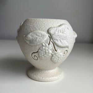 May include: A small, off-white ceramic vessel with a textured, crackled finish. The exterior features a raised, embossed design of leaves and clusters of berries. The vessel has a rounded body that tapers down to a circular pedestal base.