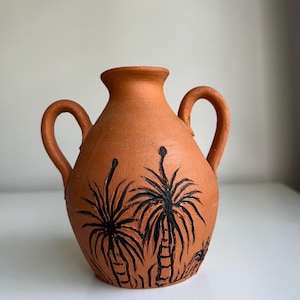 May include: A terracotta vase with two curved handles. The vase has a hand-painted design of black palm trees. The vase has a round body and a narrow neck, suitable for displaying flowers or as a decorative piece.