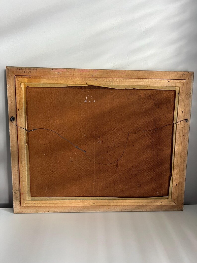 May include: Back of a framed artwork with a light brown wooden frame. The artwork's backing is a dark brown color. The frame has a wire for hanging. The upper left corner has handwritten text that reads "S. Frank-Waltz 2/16/17".
