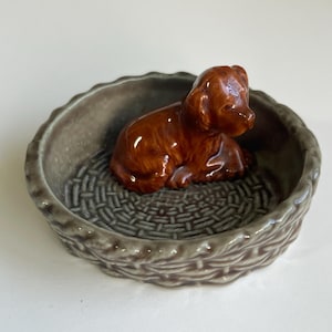 May include: A brown ceramic dog figurine in a grey, basket-style bowl. The dog is glossy and detailed, in a seated pose. The bowl has a textured interior and a woven-like exterior, creating a rustic aesthetic.