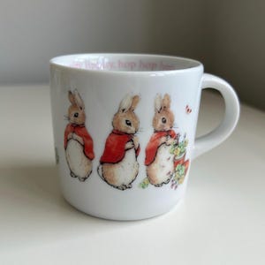 May include: A white ceramic mug featuring three illustrations of a brown rabbit wearing a red jacket. The rabbit is depicted in different poses, including one standing by a potted plant. The interior rim has the text hop hop hop printed.