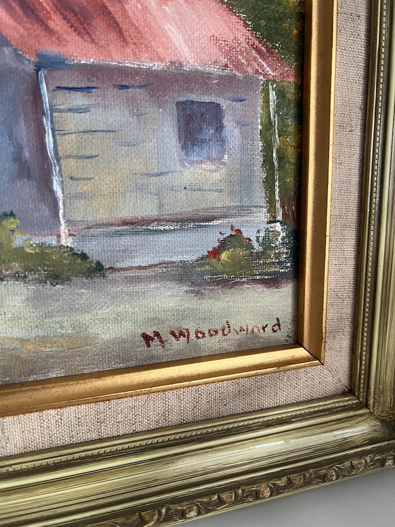 May include: An oil painting of a building with a red roof and a window, set in a landscape. The painting is signed "M. Woodward" in the lower right corner and framed in a gold-colored frame.