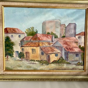 May include: Oil painting of a village scene with buildings and red roofs. The artwork is framed in a gold-colored ornate frame. The painting depicts a sunny day with a light blue sky and green trees.