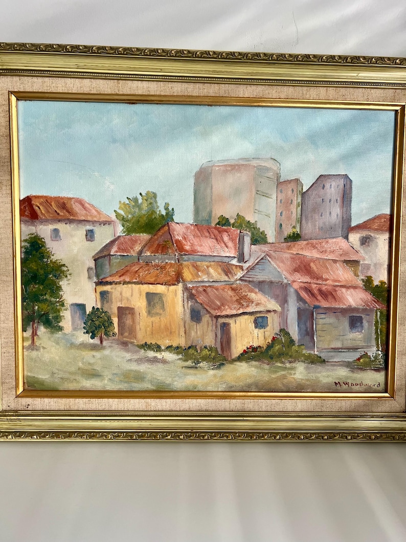 May include: Oil painting of a village scene with buildings and red roofs. The artwork is framed in a gold-colored ornate frame. The painting depicts a sunny day with a light blue sky and green trees.