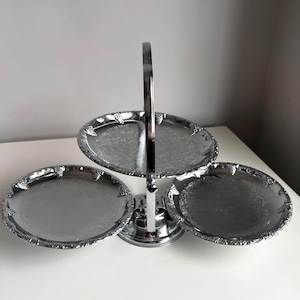May include: A silver-toned metal serving stand featuring three circular trays arranged in a tiered design. Each tray has a decorative embossed border with a grape motif and a central handle for carrying, finished with a polished metallic sheen.