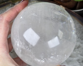 May include: A clear quartz crystal sphere, held in a hand. The sphere is transparent with internal inclusions and reflects light. The crystal ball is a popular item for home decor and spiritual practices.