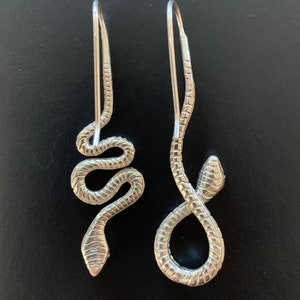 Op de afbeelding: Een paar zilveren slangen oorbellen. De oorbellen zijn gemaakt van een dunne, gewikkelde draad die in de vorm van een slang is gevormd. De slangen zijn ongeveer 5 cm lang en hebben een delicate, vrouwelijke uitstraling.