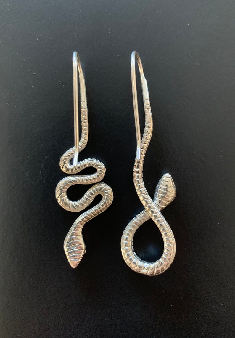 Op de afbeelding: Een paar zilveren slangen oorbellen. De oorbellen zijn gemaakt van een dunne, gewikkelde draad die in de vorm van een slang is gevormd. De slangen zijn ongeveer 5 cm lang en hebben een delicate, vrouwelijke uitstraling.