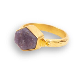 Könnte beinhalten: Ein goldener Ring mit einem sechseckigen, violetten Edelstein, der in einer goldenen Fassung sitzt. Die Ringschiene ist texturiert und hat einen kleinen goldenen Akzent.