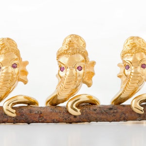 May include: Three gold-colored elephant figurines with pink gemstone eyes are arranged on a brown branch. The figurines are detailed and have a traditional design.