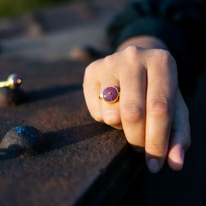 May include: A gold ring with a round, deep purple stone set in the center. The ring is on a person's finger, with a blurred background. Another ring is visible in the background.