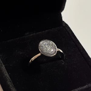 May include: A silver ring with an oval-shaped, glittery, gray stone set in a simple band. The ring is displayed in a black velvet ring box, creating a contrast between the shiny ring and the dark background.