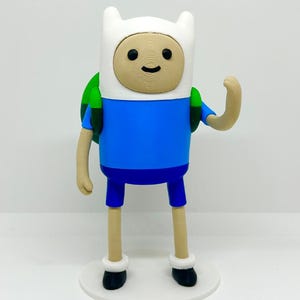Può includere: Una statuetta stampata in 3D di Finn di Adventure Time. La figura ha un cappello bianco, una maglietta blu, pantaloncini blu e uno zaino verde. La figura è su una base bianca e ha un'espressione amichevole.
