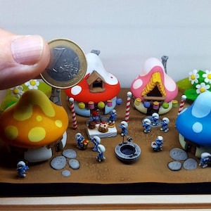 Könnte beinhalten: Eine Miniatur-Dorfszene mit vier bunten Pilzhäusern, jedes mit einem einzigartigen Design. Die Häuser sind von kleinen blauen Figuren und verschiedenen Dekorationselementen umgeben, darunter ein kleiner Brunnen und ein Kuchen.