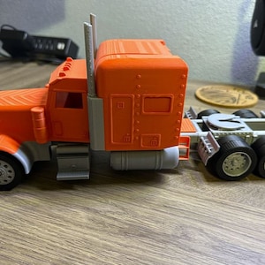 May include: A bright orange and gray toy semi-truck with a trailer hitch. The truck has a detailed cab with a windshield and side windows. The trailer is empty.