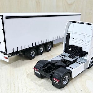 May include: White toy semi-truck and trailer with black detailing. The cab of the truck has a MAN logo on the side. The trailer is a box trailer with a rear door.