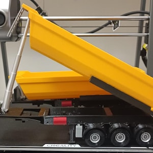 May include: A yellow dump truck model with a black chassis and a silver hydraulic lift arm. The truck bed is raised and tilted, showing the interior of the bed. The truck is on a gray platform with the word "CREATIVITY" printed on it.