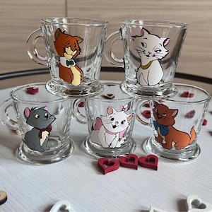 Puede incluir: Juego de seis tazas de vidrio transparente con personajes de gatos de dibujos animados. Cada taza tiene una ilustración de gato diferente en varios colores. Las tazas tienen un asa y una base. Corazones decorativos de madera están dispersos.