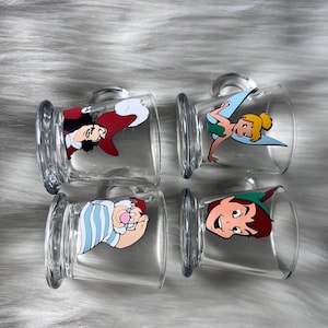 May include: Four clear glass mugs with handles, each decorated with a different character from Peter Pan. Characters include Captain Hook, Tinkerbell, Smee, and Peter Pan. The mugs are displayed on a white, fluffy surface.