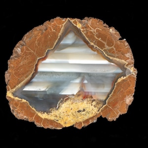 May include: A cross-section of a geode, showcasing a natural stone formation. The outer edge is a reddish-brown color with intricate crack patterns. The interior reveals layers of white and gray crystalline structures, with a small area of yellow-brown sediment.
