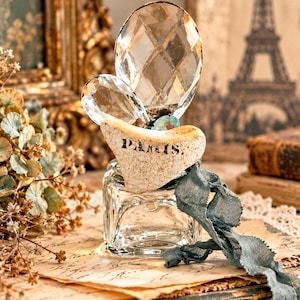 May include: A clear glass inkwell topped with a textured, off-white ceramic piece featuring the word PARIS in black lettering. Two faceted, clear crystal shapes are attached to the top, and a frayed, dusty blue fabric ribbon hangs from the side.