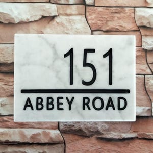 Peut inclure: Panneau de numéro de maison rectangulaire en marbre avec le numéro "151" en noir, au-dessus des mots "ABBEY ROAD". Le panneau a une bordure noire et est posé sur un mur de briques.