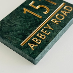 Puede incluir: Placa de dirección rectangular con un acabado de mármol verde oscuro. Los números "151" y las palabras "ABBEY ROAD" son de color dorado. Un artículo decorativo.