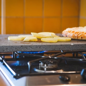 May include: A grey stone cooking surface with sliced potatoes and a cooked pizza on top, heated by a gas hob. The pizza has a crust and is topped with cheese and sauce. The kitchen has yellow tiles.