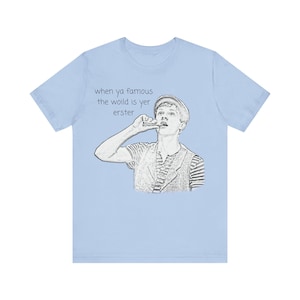 Op de afbeelding: Lichtblauw T-shirt met een zwart-wit illustratie van een man met een hoed, met een vinger voor zijn mond. De tekst op het shirt luidt "when ya famous the woild is yer erster."