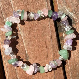 May include: A beaded bracelet composed of various polished gemstones in shades of pink, green, purple, and clear. The bracelet is arranged in a circular shape, resting on a wooden surface. The gemstones are irregularly shaped and strung together.