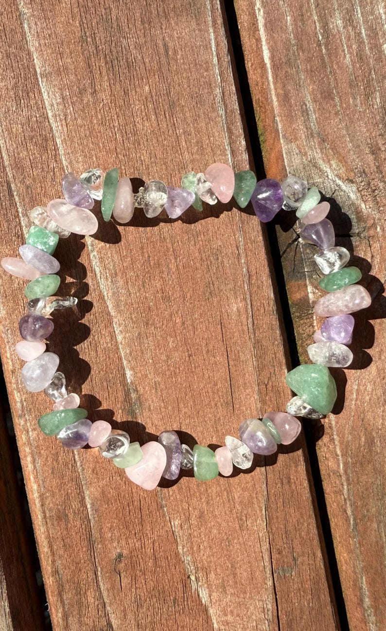 May include: A beaded bracelet composed of various polished gemstones in shades of pink, green, purple, and clear. The bracelet is arranged in a circular shape, resting on a wooden surface. The gemstones are irregularly shaped and strung together.