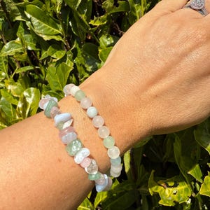 May include: Two gemstone bracelets on a wrist. One bracelet features a mix of irregular-shaped stones in pastel colors. The other bracelet has round, light pink and green beads. The background is a lush green bush.
