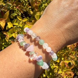 May include: Two beaded bracelets featuring a mix of colourful gemstones. The bracelets include pink, green, purple, and clear beads. The background shows blurred yellow and green foliage.