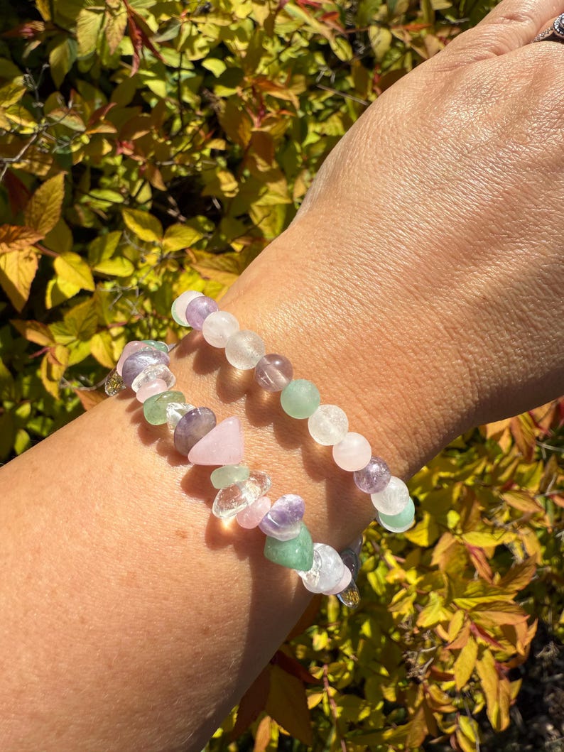 May include: Two beaded bracelets with a variety of colorful gemstones. The bracelets feature pink, green, purple, and clear beads. The background is a blurred view of yellow and green foliage.
