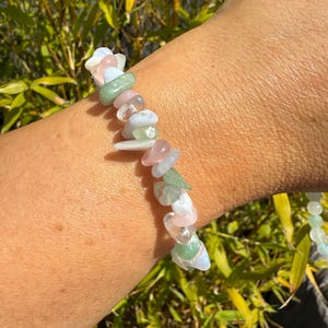 May include: A gemstone bracelet featuring a variety of colorful, irregular-shaped stones. The bracelet includes pink, green, white, and blue stones. The bracelet is worn on a wrist, with a background of green bamboo.
