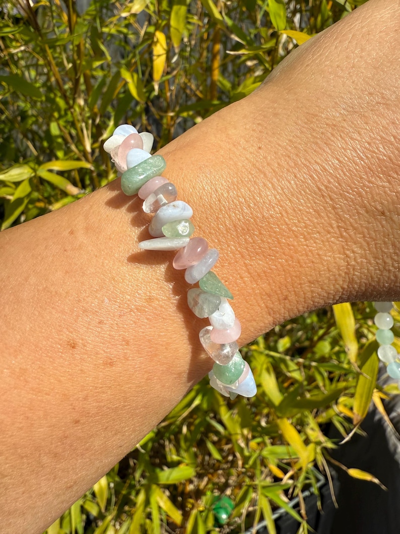 May include: A gemstone bracelet featuring a variety of colorful, irregular-shaped stones. The bracelet includes pink, green, white, and blue stones. The bracelet is worn on a wrist, with a background of green bamboo.