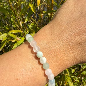 May include: A beaded bracelet featuring a mix of pastel-colored gemstones, including pink, green, and light blue. The bracelet is strung on an elastic band and is shown against a backdrop of green foliage.