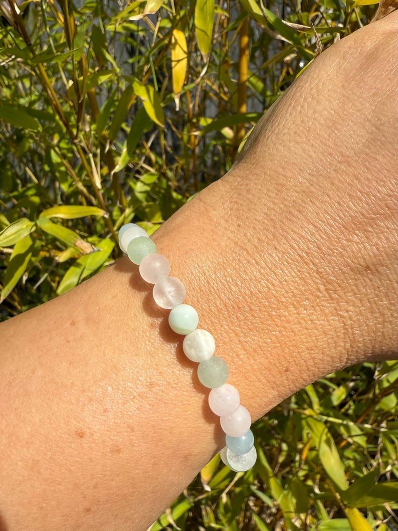 May include: A beaded bracelet featuring a mix of pastel-colored gemstones, including pink, green, and light blue. The bracelet is strung on an elastic band and is shown against a backdrop of green foliage.