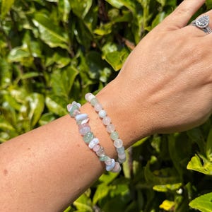 May include: Two gemstone bracelets are displayed on a wrist. One bracelet features small, round beads in shades of pink and green. The other bracelet has a mix of irregular, pastel-colored stones. The background is a lush green bush.